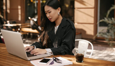 Attractive Asian Lady In Stylish Outfit Works In Computer. Brunette Tanned Woman In Black Trench Co
