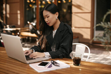 Attractive Asian lady in stylish outfit works in computer. Brunette tanned woman in black trench co